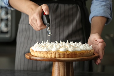 Woman toasting meringue on lemon pie with kitchen torch at table indoors, closeup