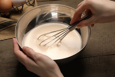 Woman whipping ingredients with balloon whisk at wooden table, closeup. Cooking delicious eggnog