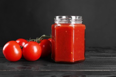 Photo of Delicious ketchup in jar and tomatoes on black wooden table. Tomato sauce