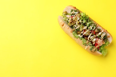 Photo of Delicious hot dog with chili peppers, lettuce, pickles and sauces on yellow background, top view. Space for text