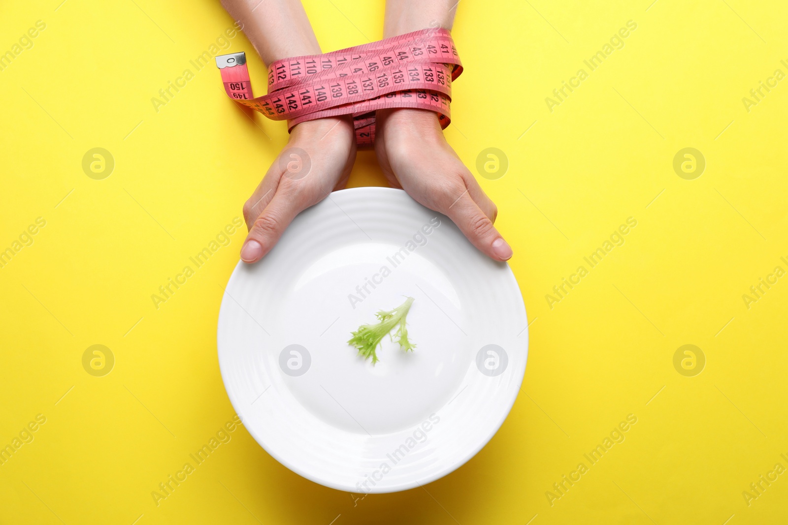 Photo of Woman holding plate with lettuce leaf in hands tied with measuring tape on yellow background, top view. Diet concept