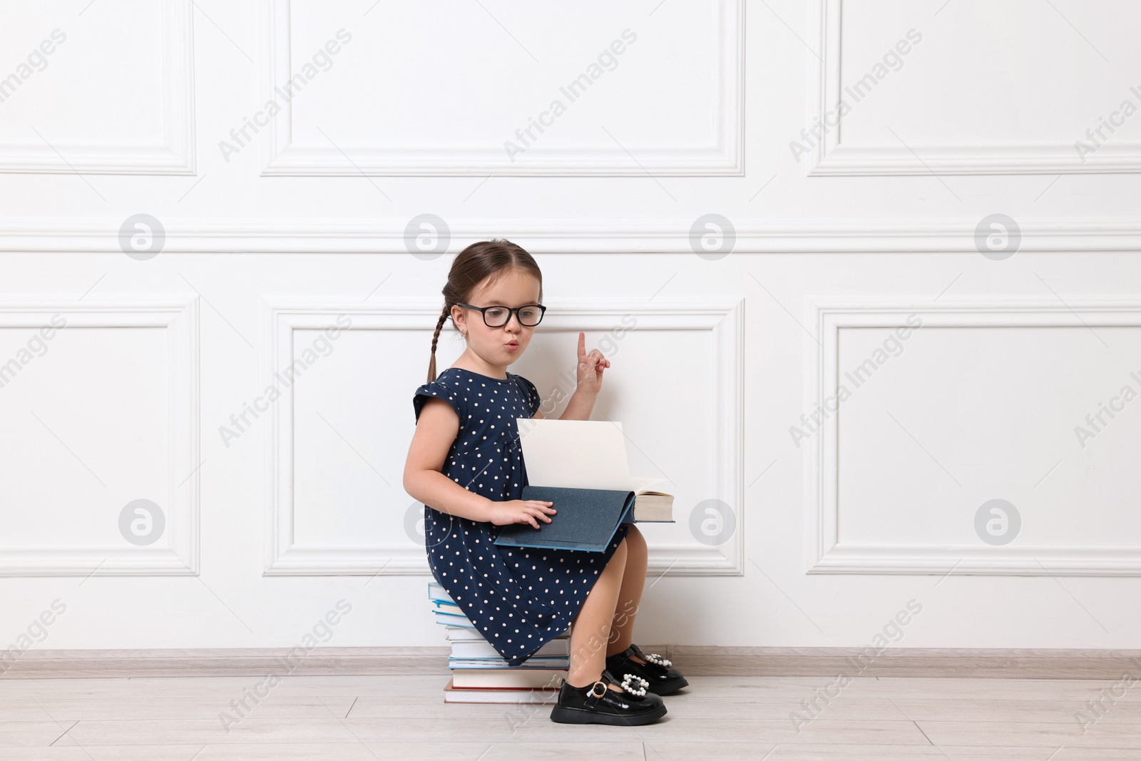 Photo of Cute little girl in glasses sitting on stack of books near white wall