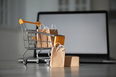 Photo of Internet shopping. Small cart with bags and box near laptop on table indoors