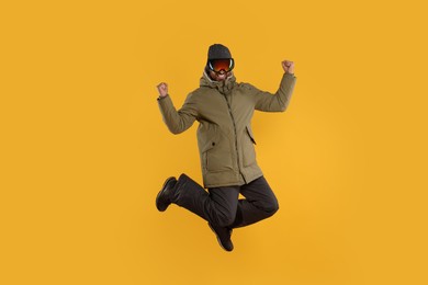Photo of Winter sports. Happy man in ski suit and goggles jumping on orange background