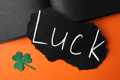 Photo of Sheet of paper with word LUCK and clover leaf on orange background, flat lay