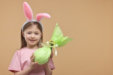 Easter celebration. Cute girl with bunny ears holding wrapped gift on beige background, space for text