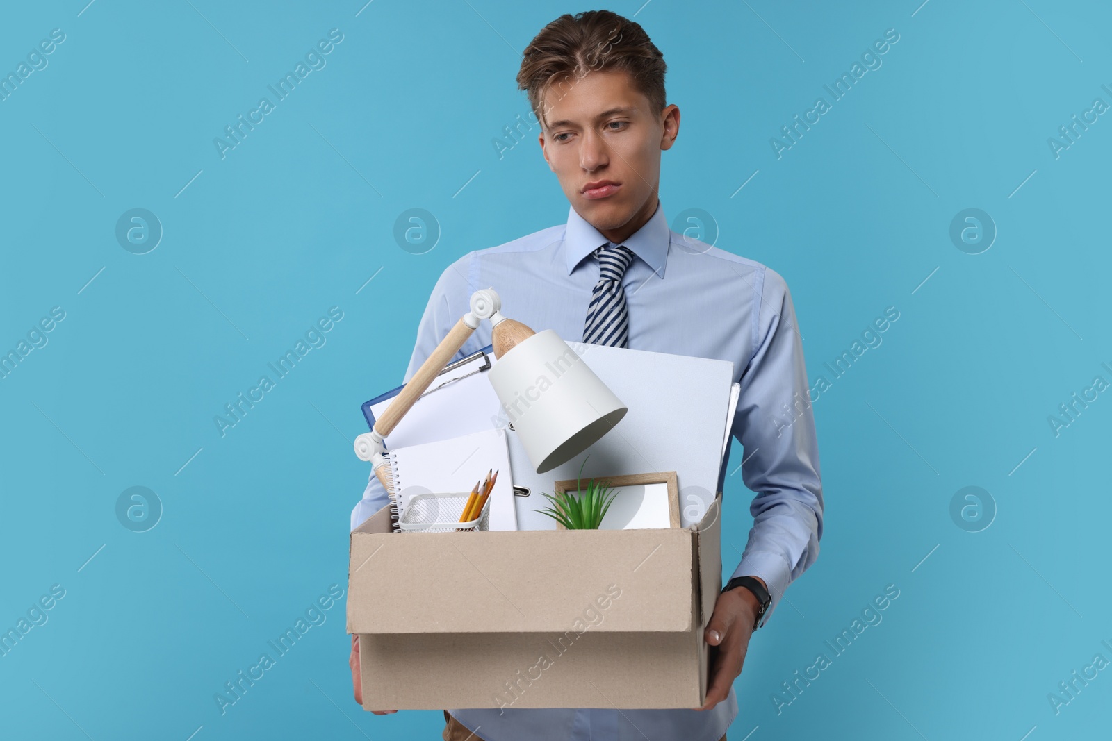 Photo of Unemployed young man with box of personal office belongings on light blue background