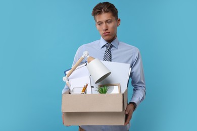 Unemployed young man with box of personal office belongings on light blue background