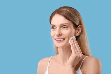 Beautiful woman removing makeup with cotton pad on light blue background, space for text