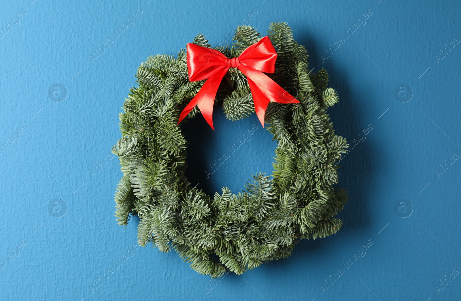 Photo of Christmas wreath made of fir tree branches with red ribbon on blue background