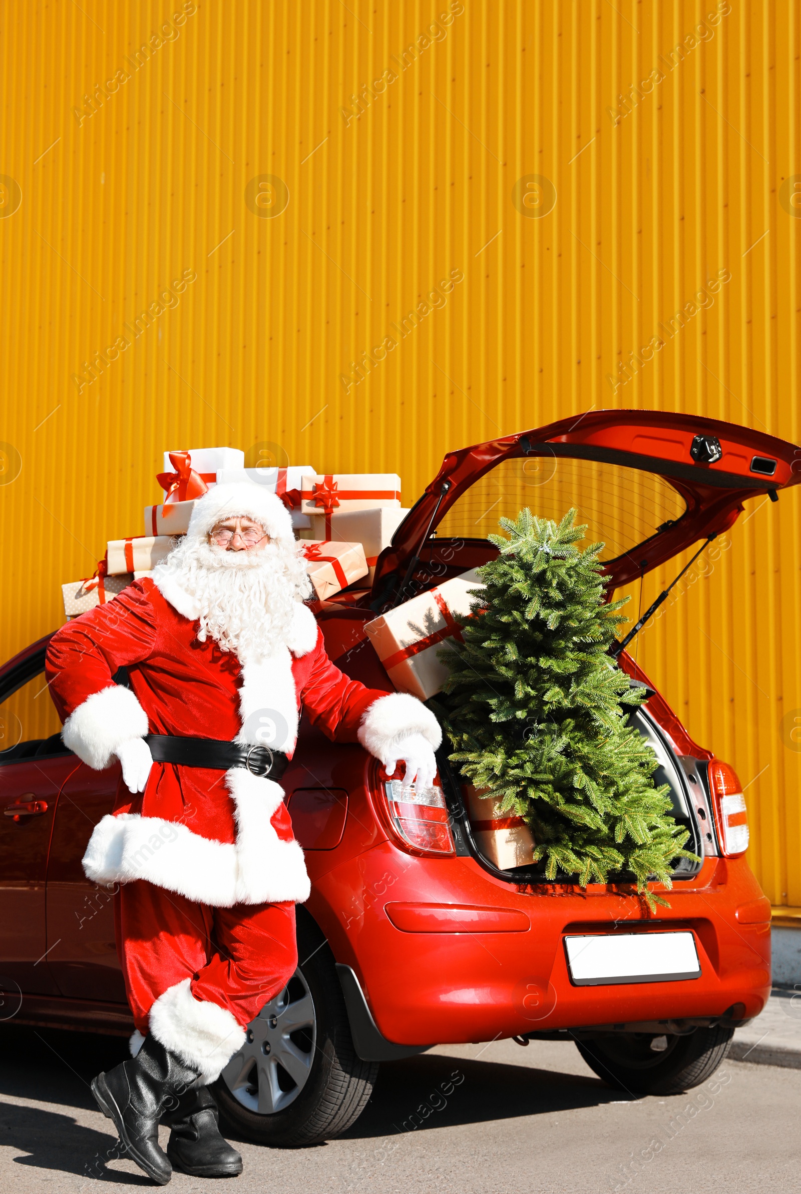 Photo of Authentic Santa Claus near red car with gift boxes and Christmas tree, outdoors