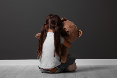 Child abuse. Upset little girl with teddy bear sitting on floor near gray wall indoors, back view