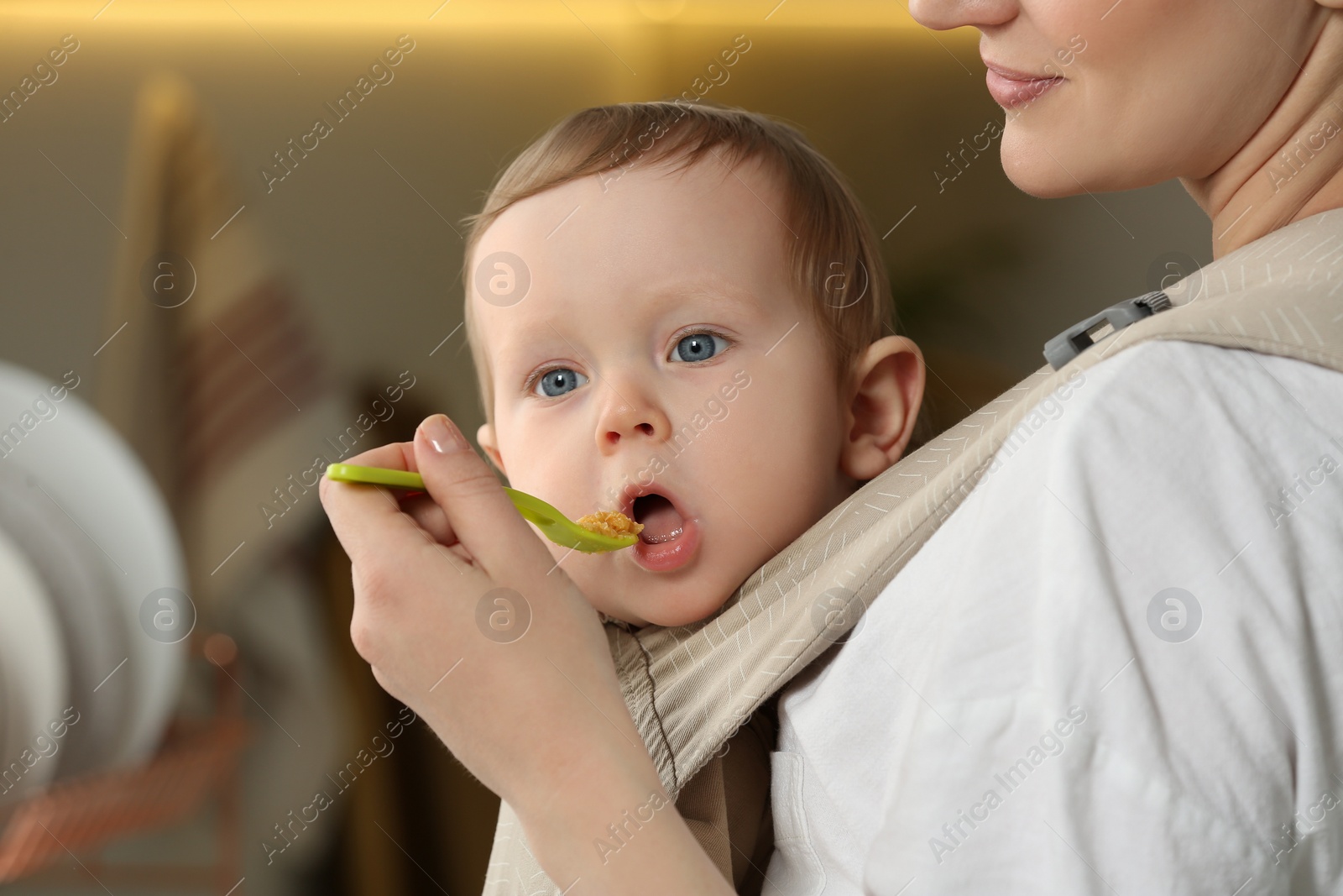 Photo of Mother feeding her cute child in sling (baby carrier) indoors