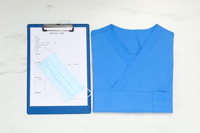 Photo of Medical uniform and clipboard on white marble table, flat lay
