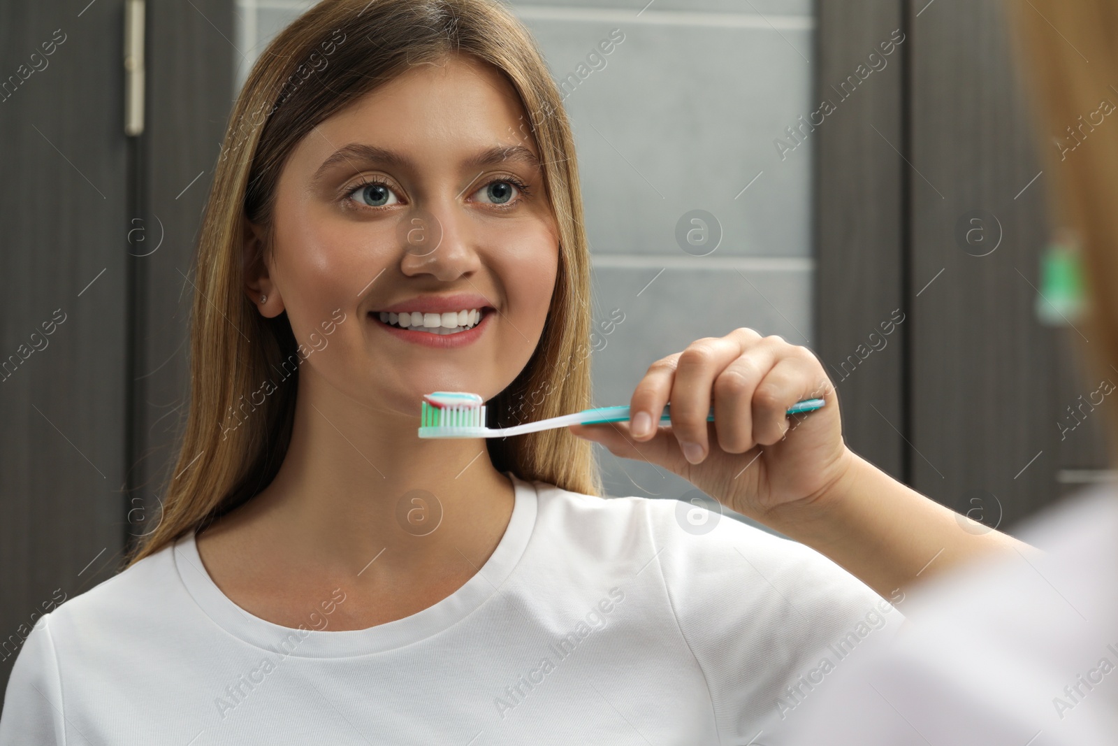 Photo of Young woman holding brush with toothpaste near mirror in bathroom