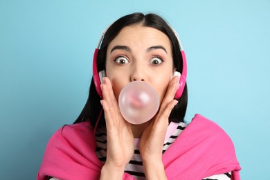 Fashionable young woman with headphones blowing bubblegum on light blue background