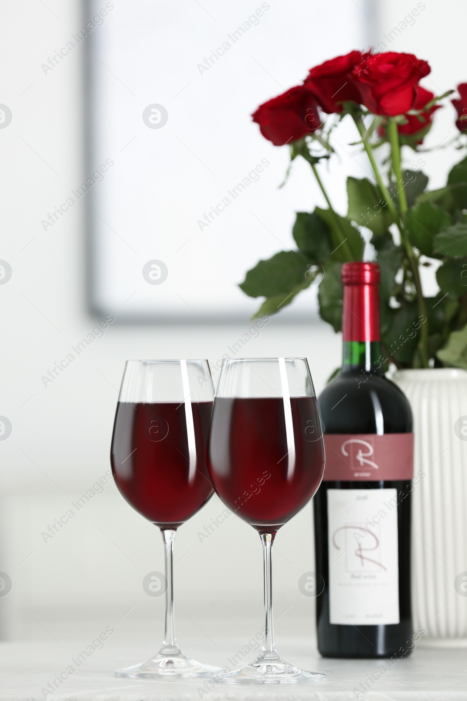 Photo of Bottle, glasses of red wine and vase with roses on table in room. Romantic date