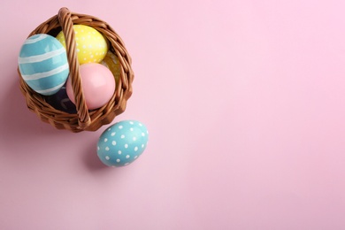 Wicker basket with painted Easter eggs on color background, top view. Space for text