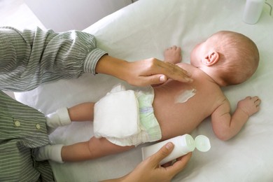 Photo of Mother applying moisturizing cream onto baby`s back on changing table indoors, above view