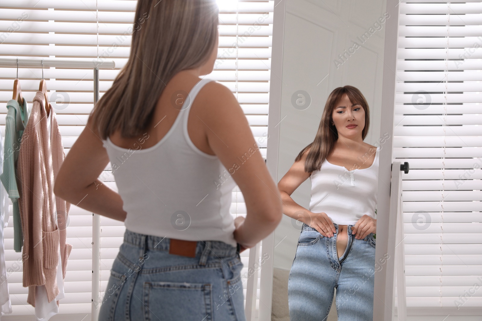 Photo of Young woman trying to put on tight jeans near mirror at home