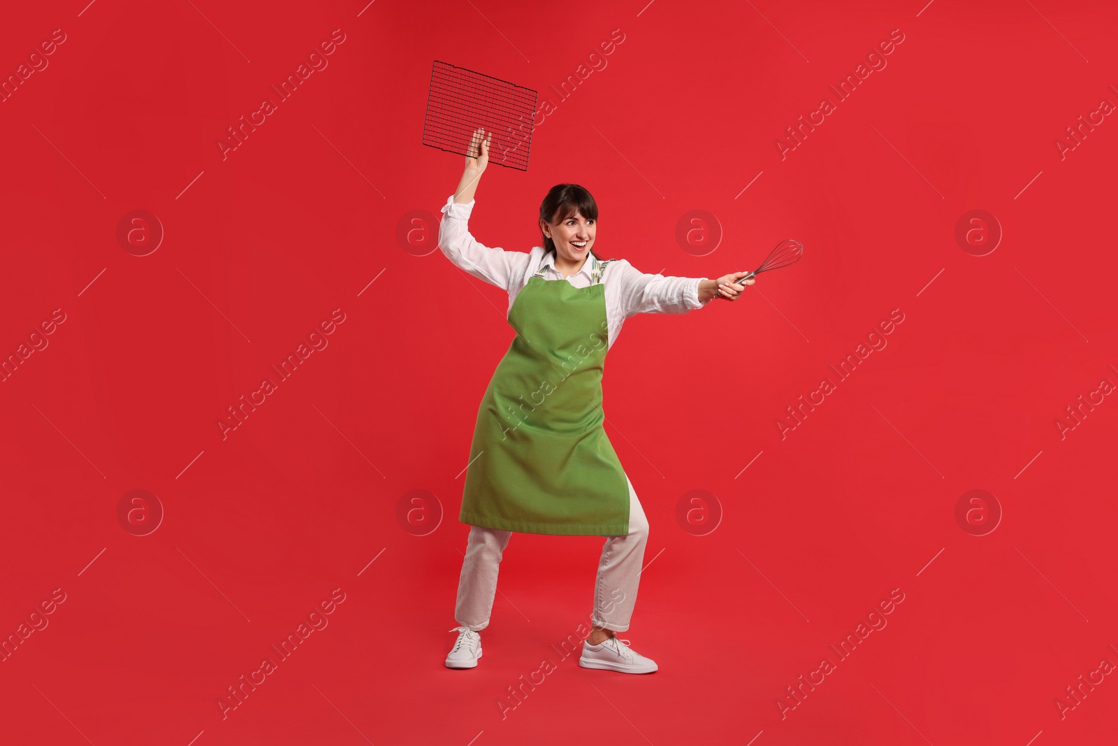 Photo of Happy confectioner in apron holding professional whisk and cooling rack on red background