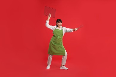 Happy confectioner in apron holding professional whisk and cooling rack on red background