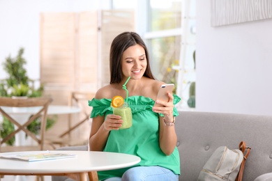 Photo of Young woman using mobile phone while drinking tasty healthy smoothie at table, indoors