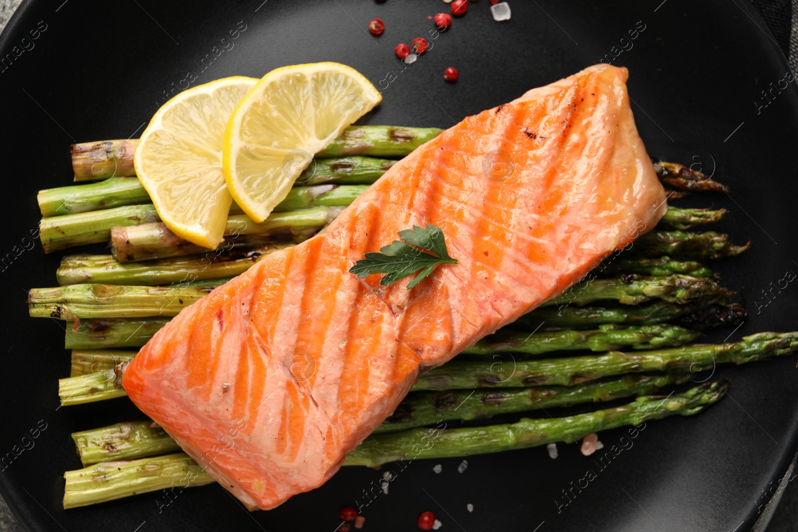 Photo of Tasty grilled salmon with asparagus, lemon and spices on black plate, top view