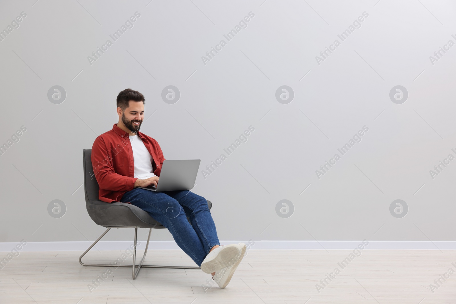 Photo of Handsome man using laptop while sitting in armchair near light grey wall indoors, space for text