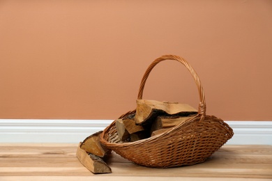 Photo of Wicker basket and firewood near brown wall indoors, space for text