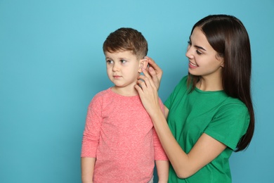 Young woman adjusting little son's hearing aid on color background