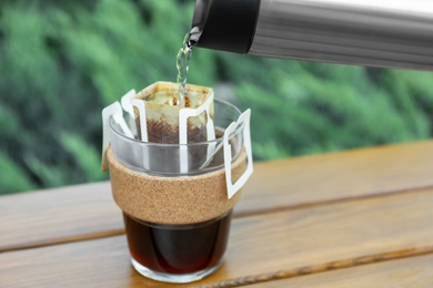 Photo of Pouring hot water into glass with drip coffee bag from thermos on wooden table, closeup