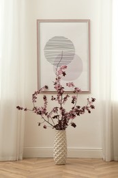 Photo of Blossoming tree twigs in vase on floor near wall with picture indoors