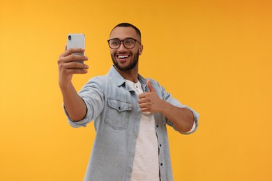 Photo of Smiling young man taking selfie with smartphone and showing thumbs up on yellow background