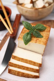 Photo of Board with smoked tofu, knife and basil on white wooden table, closeup