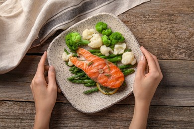 Photo of Healthy meal. Woman with plate of tasty grilled salmon and vegetables at wooden table, top view
