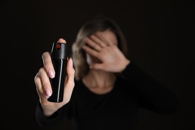 Photo of Young woman covering eyes with hand and using pepper spray on black background, focus on canister. Space for text