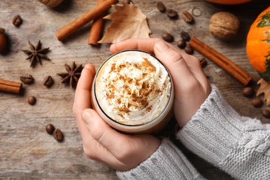 Photo of Woman holding glass of tasty pumpkin spice latte on wooden table, flat lay composition