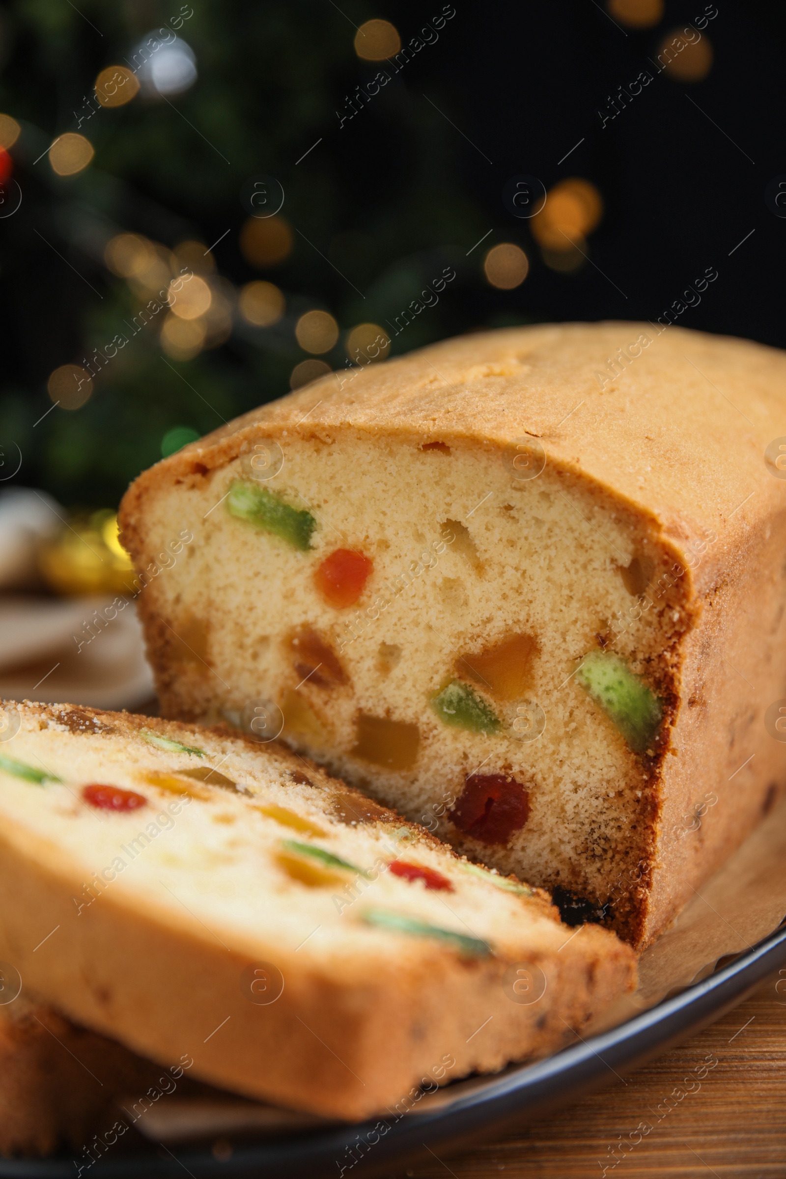 Photo of Delicious cake with candied fruits on wooden table against Christmas lights, closeup