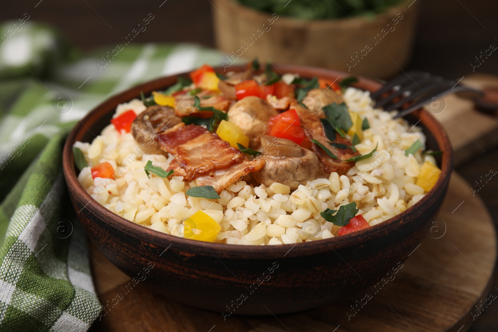 Photo of Cooked bulgur with vegetables, fried bacon and mushrooms in bowl on wooden board, closeup
