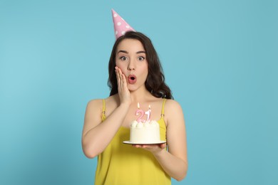 Photo of Coming of age party - 21st birthday. Surprised woman holding delicious cake with number shaped candles on light blue background