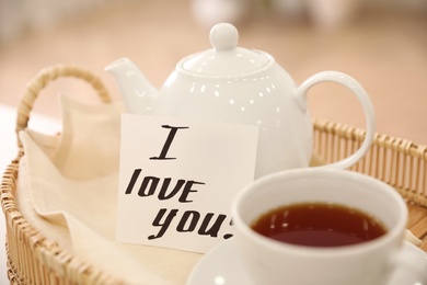 Paper note with handwritten words I Love You and tea on wicker tray, closeup