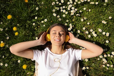 Happy woman listening to audiobook while lying on grass among blooming daisies, top view