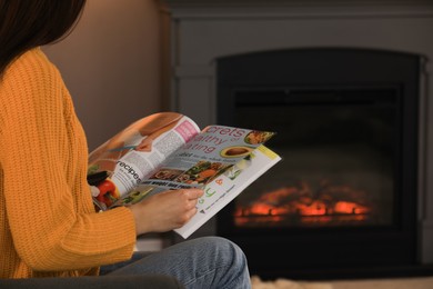 Photo of Young woman reading magazine near fireplace at home, closeup. Space for text