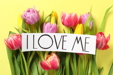 Piece of paper with handwritten phrase I Love Me  and tulips on yellow background, flat lay