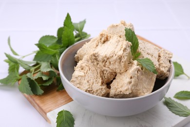 Bowl with pieces of tasty halva and mint leaves on white table, closeup