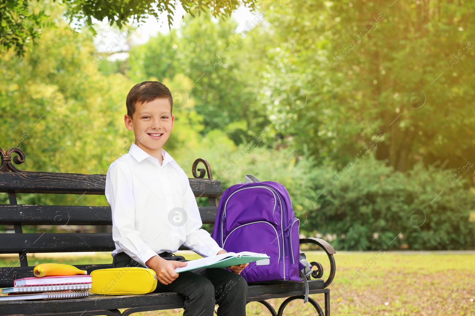 Photo of Cute little school child with stationery reading book on bench in park