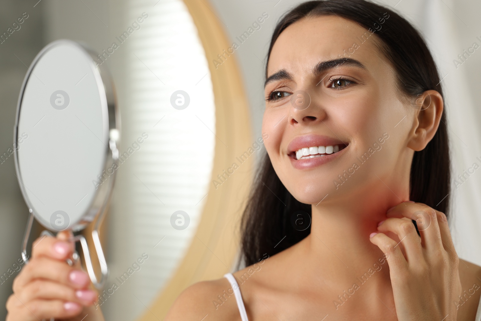 Photo of Suffering from allergy. Young woman looking in mirror and scratching her neck indoors