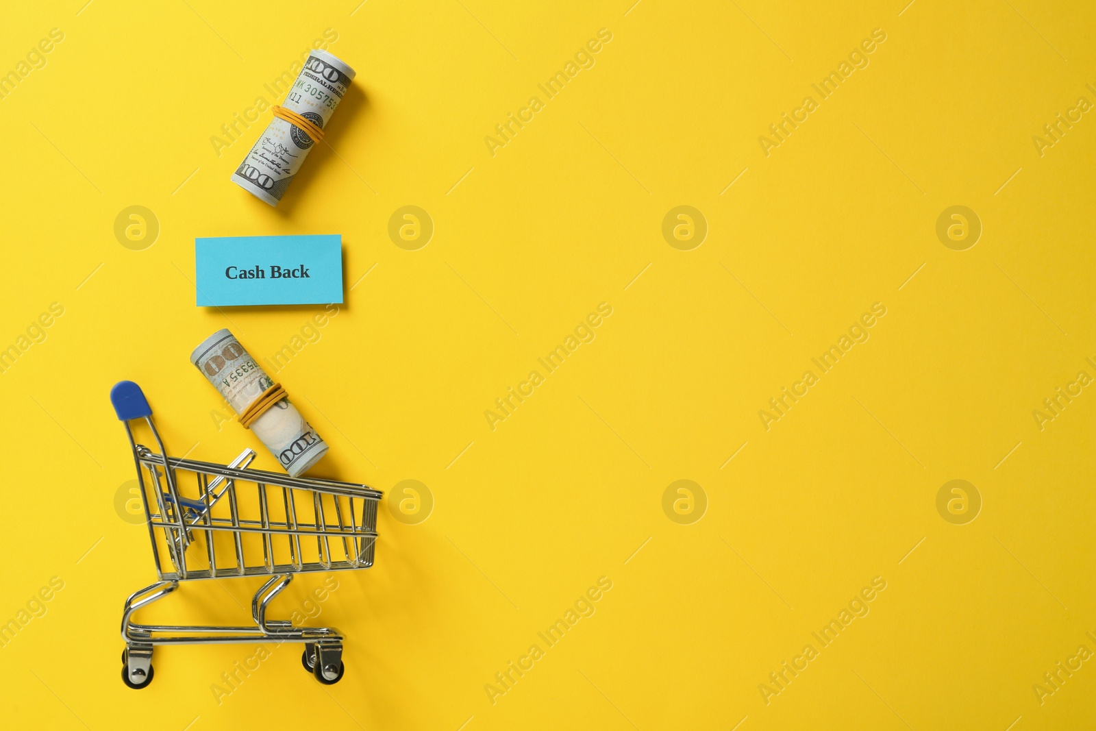 Photo of Card with word Cashback, rolled dollar banknotes and shopping cart on yellow background, flat lay. Space for text