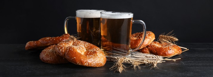 Tasty pretzels, glasses of beer and wheat spikes on black table against dark background. Banner design